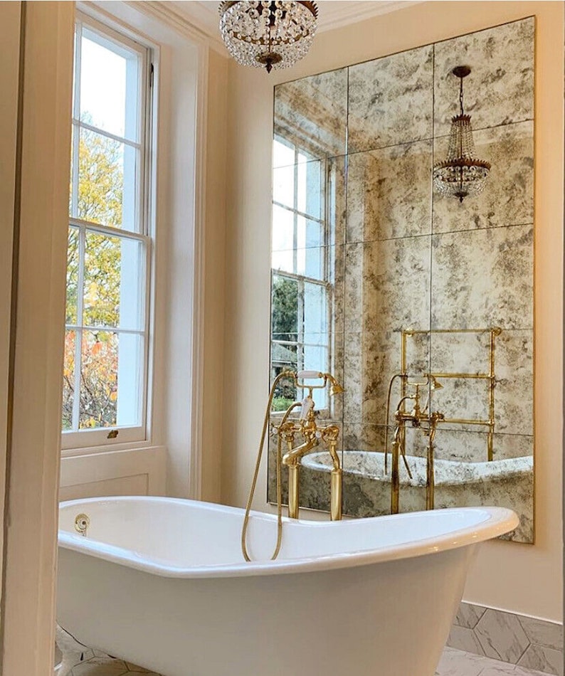 Puede incluir: Una ba&ntilde;era exenta blanca con grifer&iacute;a dorada y una ducha de mano. Detr&aacute;s de la ba&ntilde;era hay un gran espejo de pared rectangular con un acabado antiguo y moteado, que refleja una l&aacute;mpara de ara&ntilde;a de cristal y un toallero dorado.