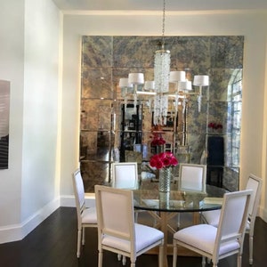 May include: A dining room set featuring a round glass-topped table with a wooden base, surrounded by four white upholstered chairs. A crystal chandelier hangs above, and a large wall-mounted antique-style grid mirror reflects the room.