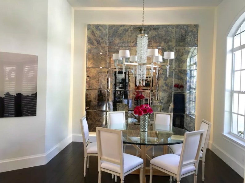 May include: A dining room set featuring a round glass-topped table with a wooden base, surrounded by four white upholstered chairs. A crystal chandelier hangs above, and a large wall-mounted antique-style grid mirror reflects the room.