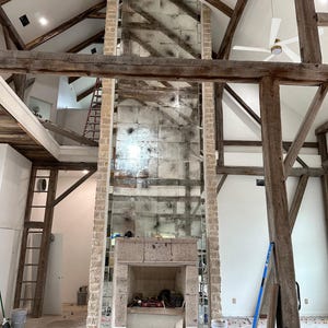 May include: A tall fireplace with a stone surround and a large, rectangular, antiqued mirror panel extending to the vaulted ceiling. The structure features exposed wooden beams, white walls, and a herringbone floor with red tile spacers.