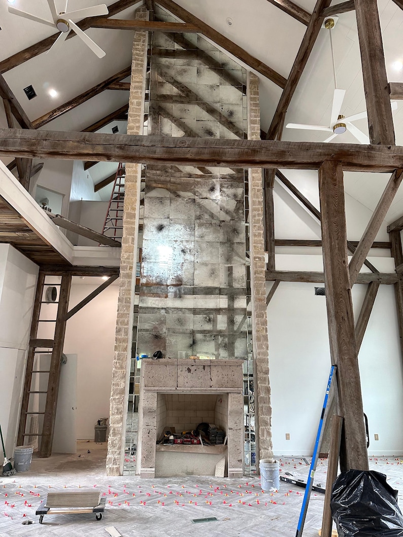 May include: A tall fireplace with a stone surround and a large, rectangular, antiqued mirror panel extending to the vaulted ceiling. The structure features exposed wooden beams, white walls, and a herringbone floor with red tile spacers.