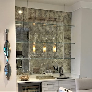 May include: A home bar setup featuring three horizontal glass shelves suspended by thin metal rods against an antique-style distressed mirror backsplash. The shelves hold clear wine glasses, while the white countertop includes a sink and a bowl of corks.