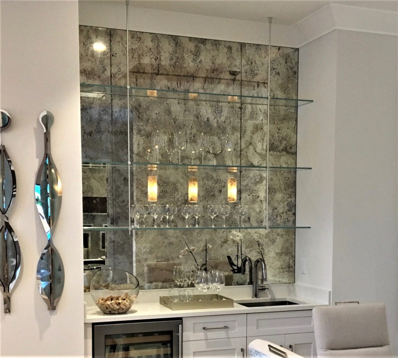 May include: A home bar setup featuring three horizontal glass shelves suspended by thin metal rods against an antique-style distressed mirror backsplash. The shelves hold clear wine glasses, while the white countertop includes a sink and a bowl of corks.