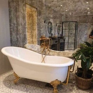 May include: A white clawfoot bathtub with gold-toned feet and a matching gold-toned faucet fixture. The tub is positioned in a room with patterned floor tiles and a large wall mirror reflecting a bathroom vanity and a black-framed shower enclosure.