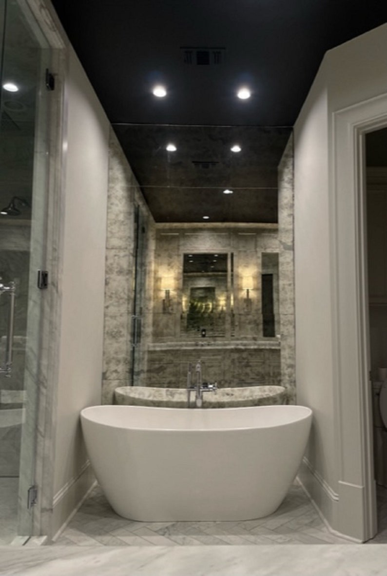 Puede incluir: Una ba&ntilde;era exenta ovalada de color blanco con un grifo cromado centrado en el borde. La ba&ntilde;era est&aacute; situada contra una pared cubierta con grandes paneles de espejo de acabado antiguo y desgastado que reflejan el ba&ntilde;o y las luces del techo.