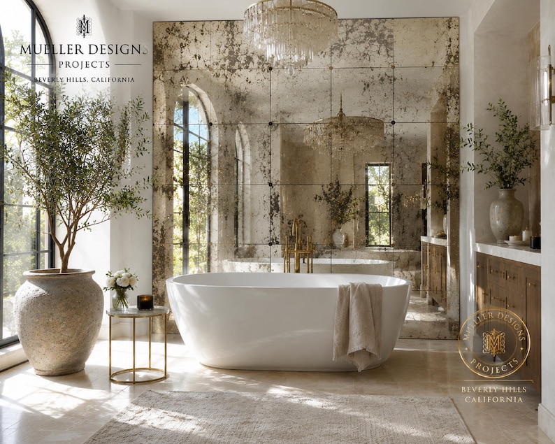 May include: A white freestanding oval bathtub with a beige towel draped over the side, positioned in front of a large antique-style grid mirror. The room features a tall olive tree in a textured stone planter, a small gold side table, and a chandelier.