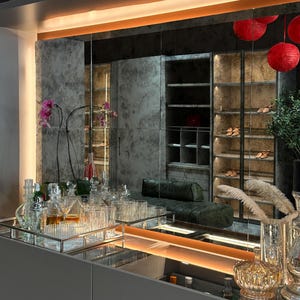 May include: A modern bar cabinet featuring a sleek gray surface with a rectangular silver tray holding various clear glass decanters and tumblers. A large mirror reflects a walk-in closet with illuminated shelving, a green leather bench, and red lanterns.