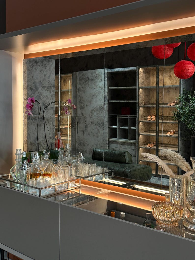 May include: A modern bar cabinet featuring a sleek gray surface with a rectangular silver tray holding various clear glass decanters and tumblers. A large mirror reflects a walk-in closet with illuminated shelving, a green leather bench, and red lanterns.