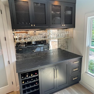 May include: A black home bar cabinet featuring upper cabinets with mesh panels, a mirrored backsplash, and gold hardware. The unit includes a black countertop, a built-in wine rack, two lower cabinet doors, and three drawers with gold handles.