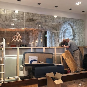 May include: A large wall installation featuring a grid of rectangular antique-style mirrors with a distressed, mottled silver finish. The mirrors reflect a room with arched architectural details and a multi-tiered light fixture above a wooden frame.