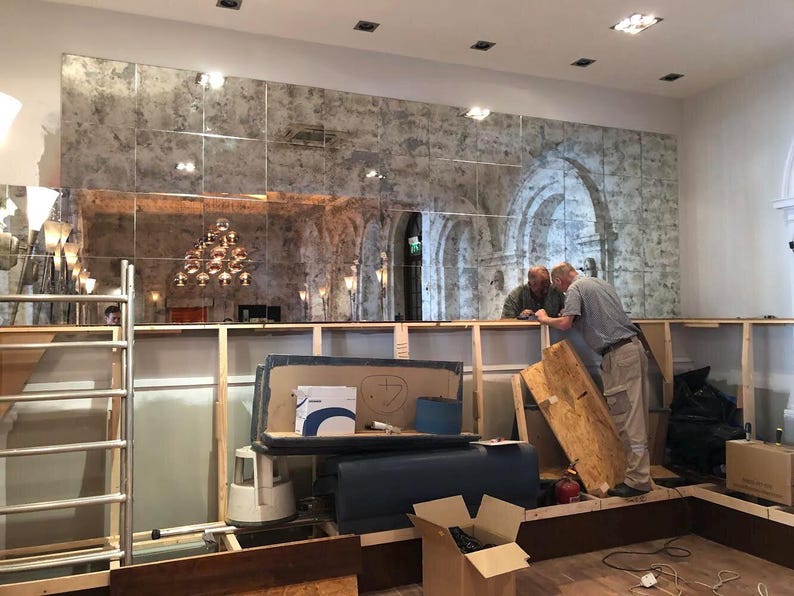 May include: A large wall installation featuring a grid of rectangular antique-style mirrors with a distressed, mottled silver finish. The mirrors reflect a room with arched architectural details and a multi-tiered light fixture above a wooden frame.