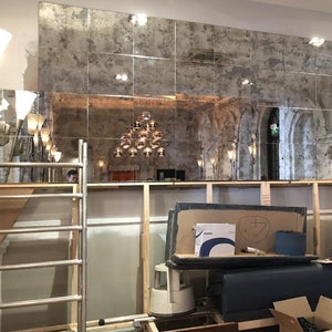 Puede incluir: Una gran instalaci&oacute;n de pared compuesta por azulejos de espejo cuadrados y envejecidos con un acabado moteado y desgastado. Los espejos reflejan una habitaci&oacute;n con una l&aacute;mpara colgante de varias bombillas, arcos arquitect&oacute;nicos y una obra.