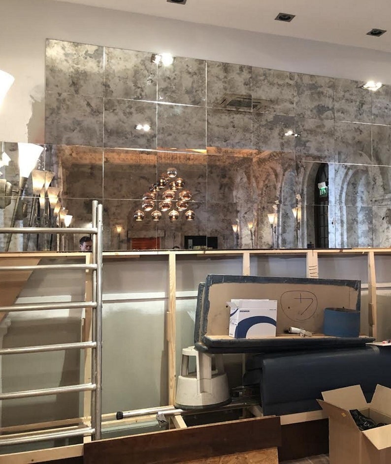 Puede incluir: Una gran instalaci&oacute;n de pared compuesta por azulejos de espejo cuadrados y envejecidos con un acabado moteado y desgastado. Los espejos reflejan una habitaci&oacute;n con una l&aacute;mpara colgante de varias bombillas, arcos arquitect&oacute;nicos y una obra.