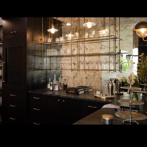May include: A home bar setup featuring a dark cabinetry base with gold hardware, a black stone countertop, and a suspended brass shelving unit holding various clear glassware. A mirrored backsplash reflects the room, and a three-tier metal stand sits nearby.
