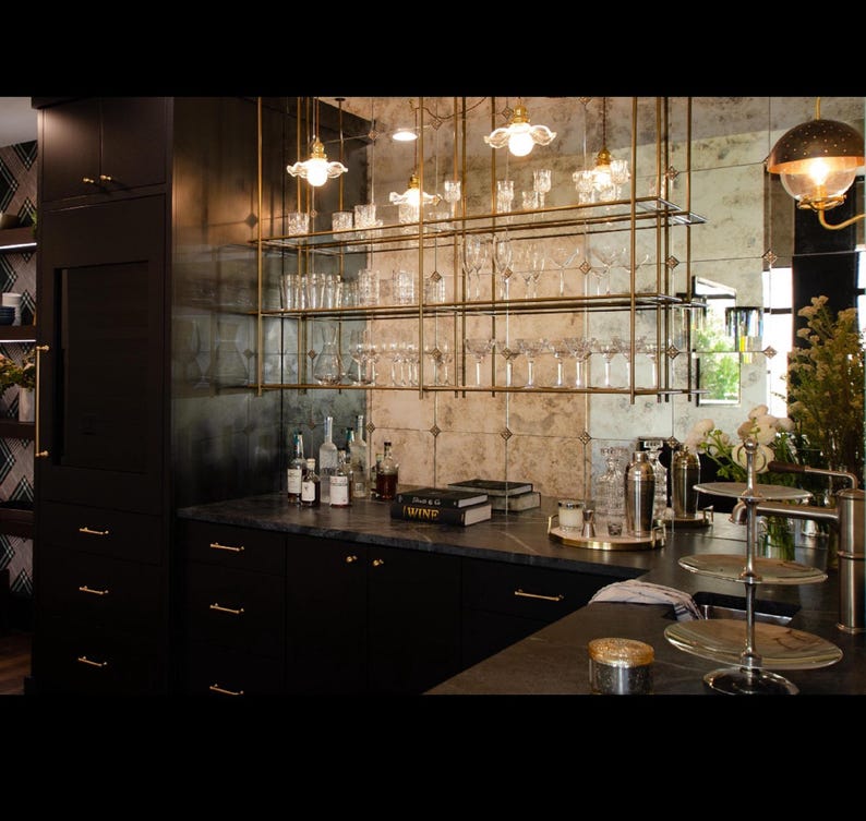 May include: A home bar setup featuring a dark cabinetry base with gold hardware, a black stone countertop, and a suspended brass shelving unit holding various clear glassware. A mirrored backsplash reflects the room, and a three-tier metal stand sits nearby.