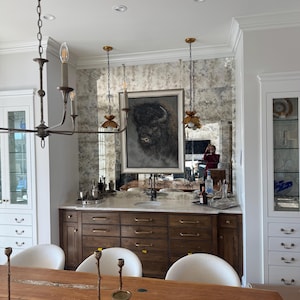 May include: A wooden bar cabinet with a marble countertop, featuring a framed bison portrait hanging on an antique-style mirror backsplash. The unit includes multiple drawers with gold-toned hardware, flanked by white cabinetry and a dining area.