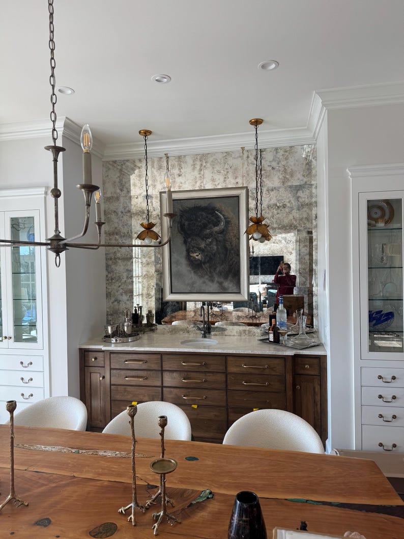 May include: A wooden bar cabinet with a marble countertop, featuring a framed bison portrait hanging on an antique-style mirror backsplash. The unit includes multiple drawers with gold-toned hardware, flanked by white cabinetry and a dining area.