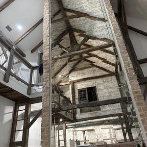 May include: A tall, rectangular floor-to-ceiling mirror panel installed against a stone fireplace chimney. The mirror reflects a high vaulted ceiling with exposed dark wooden beams, a lofted balcony area, and white textured walls.