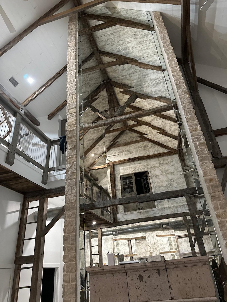 May include: A tall, rectangular floor-to-ceiling mirror panel installed against a stone fireplace chimney. The mirror reflects a high vaulted ceiling with exposed dark wooden beams, a lofted balcony area, and white textured walls.