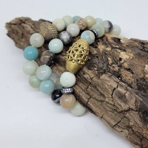 May include: Two beaded bracelets featuring a combination of light blue, white and brown beads. One bracelet has a gold metal charm with a geometric design. The bracelets are displayed on a piece of driftwood.