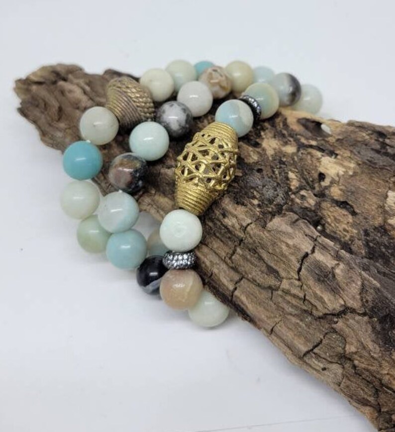 May include: Two beaded bracelets featuring a combination of light blue, white and brown beads. One bracelet has a gold metal charm with a geometric design. The bracelets are displayed on a piece of driftwood.