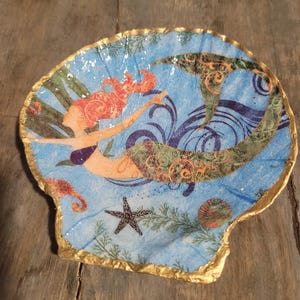 May include: A seashell-shaped decorative dish with a mermaid design. The dish features a blue background with a mermaid illustration in orange, green, and purple. The rim is trimmed with gold paint. Other sea creatures are also depicted.