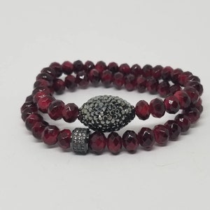 Pode incluir: Um bracelete de duas voltas feito de contas facetadas vermelho-escuro com um fecho oval preto adornado com pequenos strass transparentes.