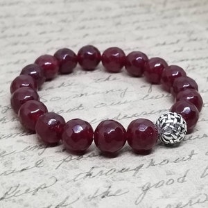 May include: A red gemstone bracelet with a silver charm. The bracelet is made of round beads and has a silver charm in the shape of a woven ball.