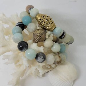 May include: Three bracelets made of light blue, white and brown beads with gold accents. The bracelets are stacked on top of each other and are sitting on a white coral-like surface.