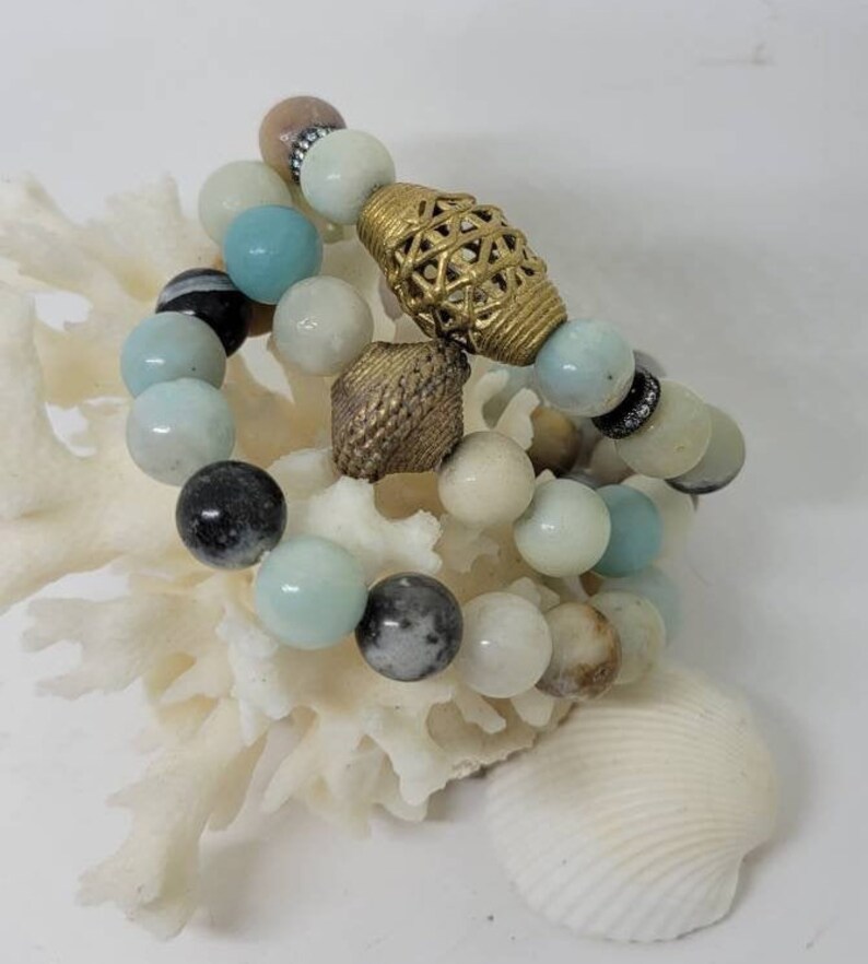 May include: Three bracelets made of light blue, white and brown beads with gold accents. The bracelets are stacked on top of each other and are sitting on a white coral-like surface.