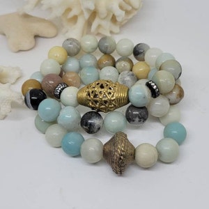 May include: Three layered bracelets with light blue, white and brown beads. The top bracelet has a gold metal bead with a lattice design. The middle bracelet has a gold metal bead with a diamond-shaped design. The bottom bracelet has a gold metal bead with a cone-shaped design.