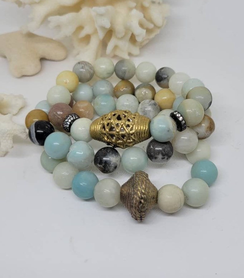 May include: Three layered bracelets with light blue, white and brown beads. The top bracelet has a gold metal bead with a lattice design. The middle bracelet has a gold metal bead with a diamond-shaped design. The bottom bracelet has a gold metal bead with a cone-shaped design.