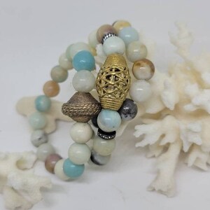 May include: Three layered bracelets made of light blue, white and brown beads with gold accents. The bracelets are stacked on top of each other and are resting on a white coral.