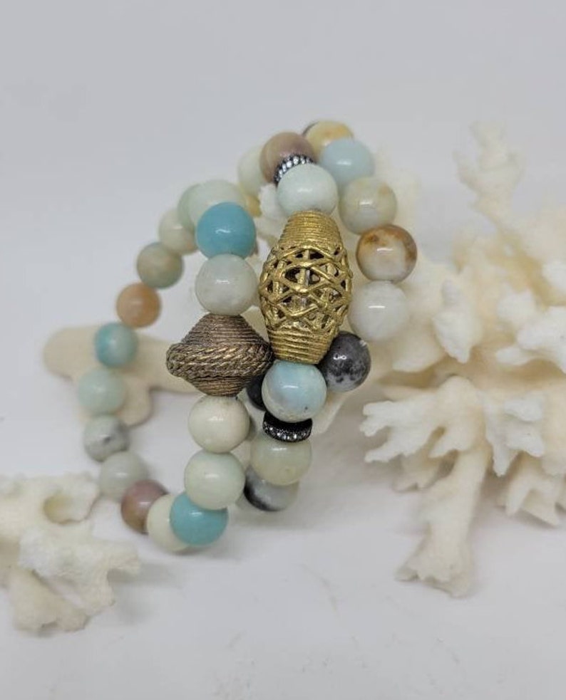 May include: Three layered bracelets made of light blue, white and brown beads with gold accents. The bracelets are stacked on top of each other and are resting on a white coral.