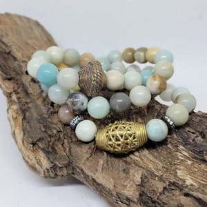 May include: Four stacked bracelets made of light blue, green, and brown beads with gold accents. The bracelets are on a piece of driftwood.