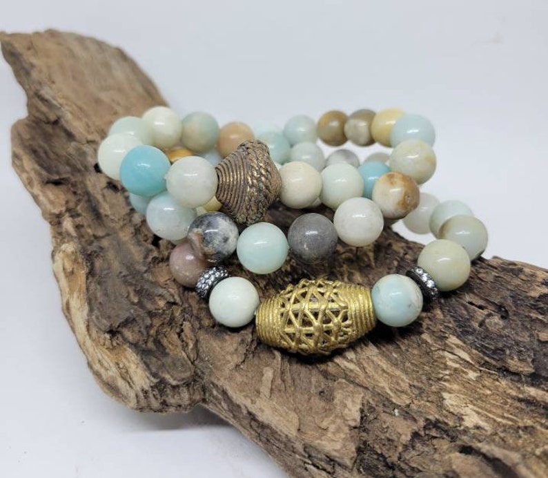 May include: Four stacked bracelets made of light blue, green, and brown beads with gold accents. The bracelets are on a piece of driftwood.