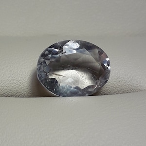 May include: A clear oval-shaped gemstone with a faceted cut. The gemstone is on a white background.