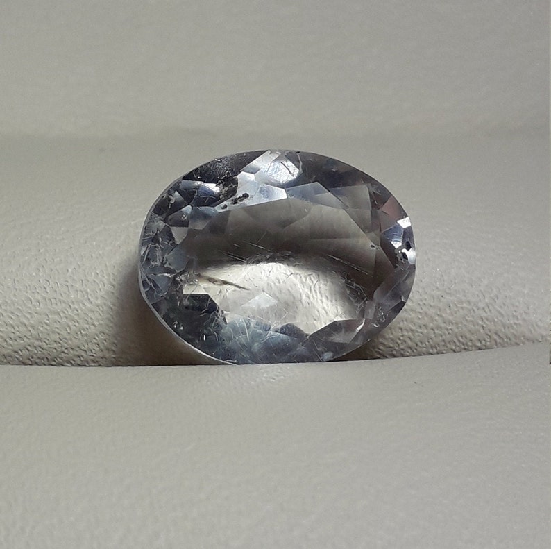 May include: A clear oval-shaped gemstone with a faceted cut. The gemstone is on a white background.