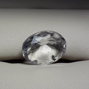 May include: A clear, faceted oval-shaped gemstone with a slight gray hue. The gemstone is resting on a white surface.