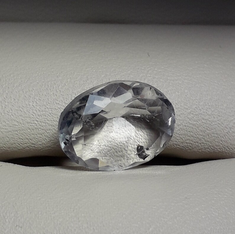 May include: A clear, faceted oval-shaped gemstone with a slight gray hue. The gemstone is resting on a white surface.