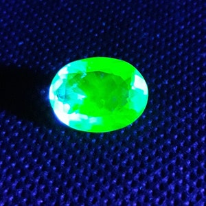 May include: A bright green oval-shaped gemstone with a faceted cut, displayed on a blue textured surface.