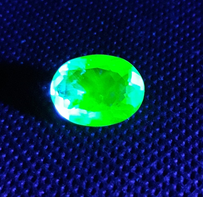 May include: A bright green oval-shaped gemstone with a faceted cut, displayed on a blue textured surface.