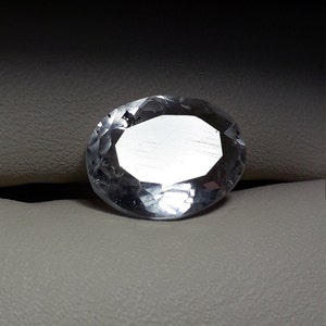 May include: A clear, oval-shaped gemstone with a faceted cut. The gemstone is set on a white surface.