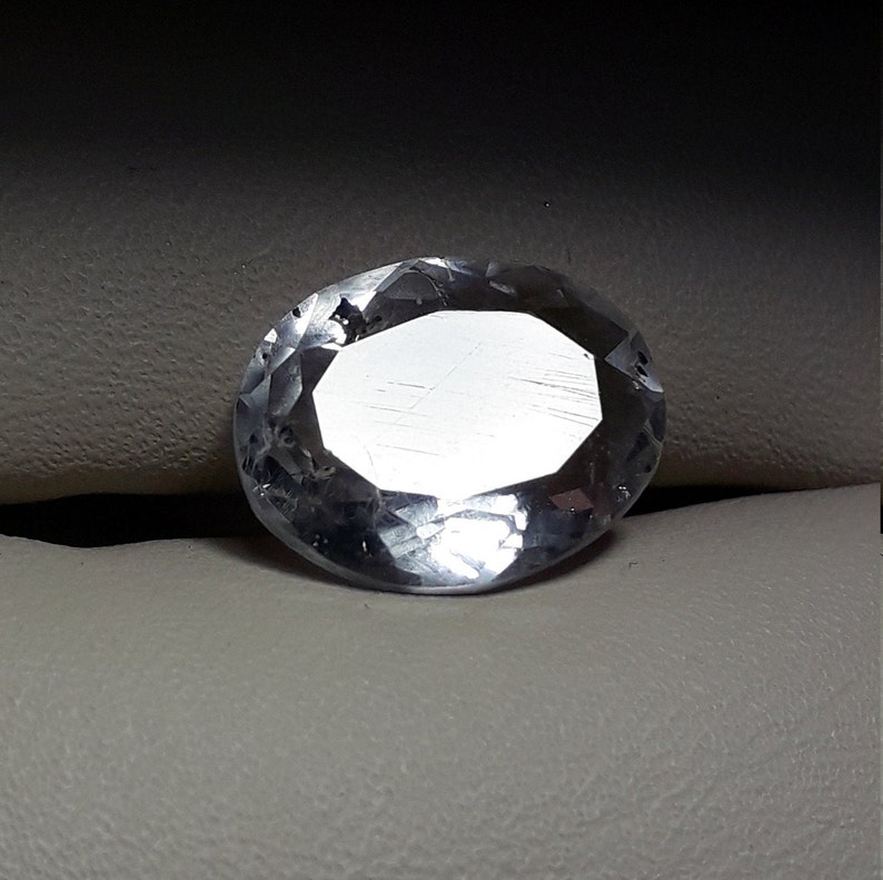 May include: A clear, oval-shaped gemstone with a faceted cut. The gemstone is set on a white surface.