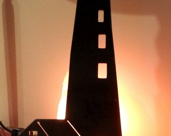May include: A black metal silhouette of a lighthouse with a small attached building, mounted on a rectangular wooden base. The structure features cut-out windows and a lantern room, illuminated from behind by a warm, glowing orange light source.
