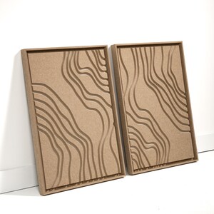 May include: Two rectangular wall art pieces with a topographic design. The art features a series of curved, parallel lines carved into a light brown surface, set within a matching wooden frame. The artwork is leaning against a white wall.