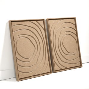 May include: Two rectangular, light brown wooden wall art panels. Each panel features a series of concentric, curved lines, creating a topographic or wood grain effect. The panels are leaning against a white wall.