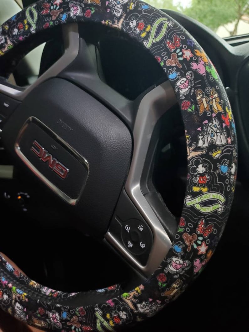 Puede incluir: Funda de volante negra con un estampado de Disney colorido que presenta a Mickey Mouse, Minnie Mouse, Donald Duck, Goofy y otros personajes de Disney. El estampado tambi&eacute;n incluye las palabras "Walt Disney World" y "Disneyland".