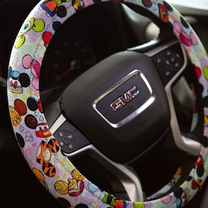May include: A colorful steering wheel cover with a Disney character theme. The cover features Mickey Mouse, Minnie Mouse, Stitch, and other Disney characters on a white background with pink, blue, and yellow accents.