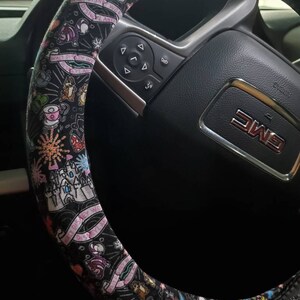 Puede incluir: Funda de volante negra con un estampado de Disney colorido que presenta personajes como Mickey Mouse, Minnie Mouse y el castillo de Cenicienta. El texto "Walt Disney World" est&aacute; impreso en la funda.